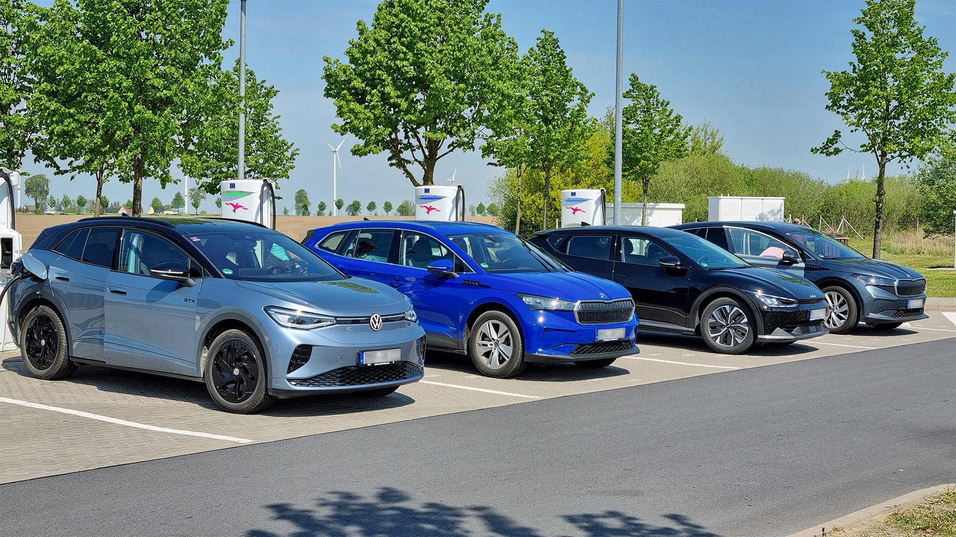 Four electric cars from different manufacturers lined up at a rapid-charging hub beside a motorway.