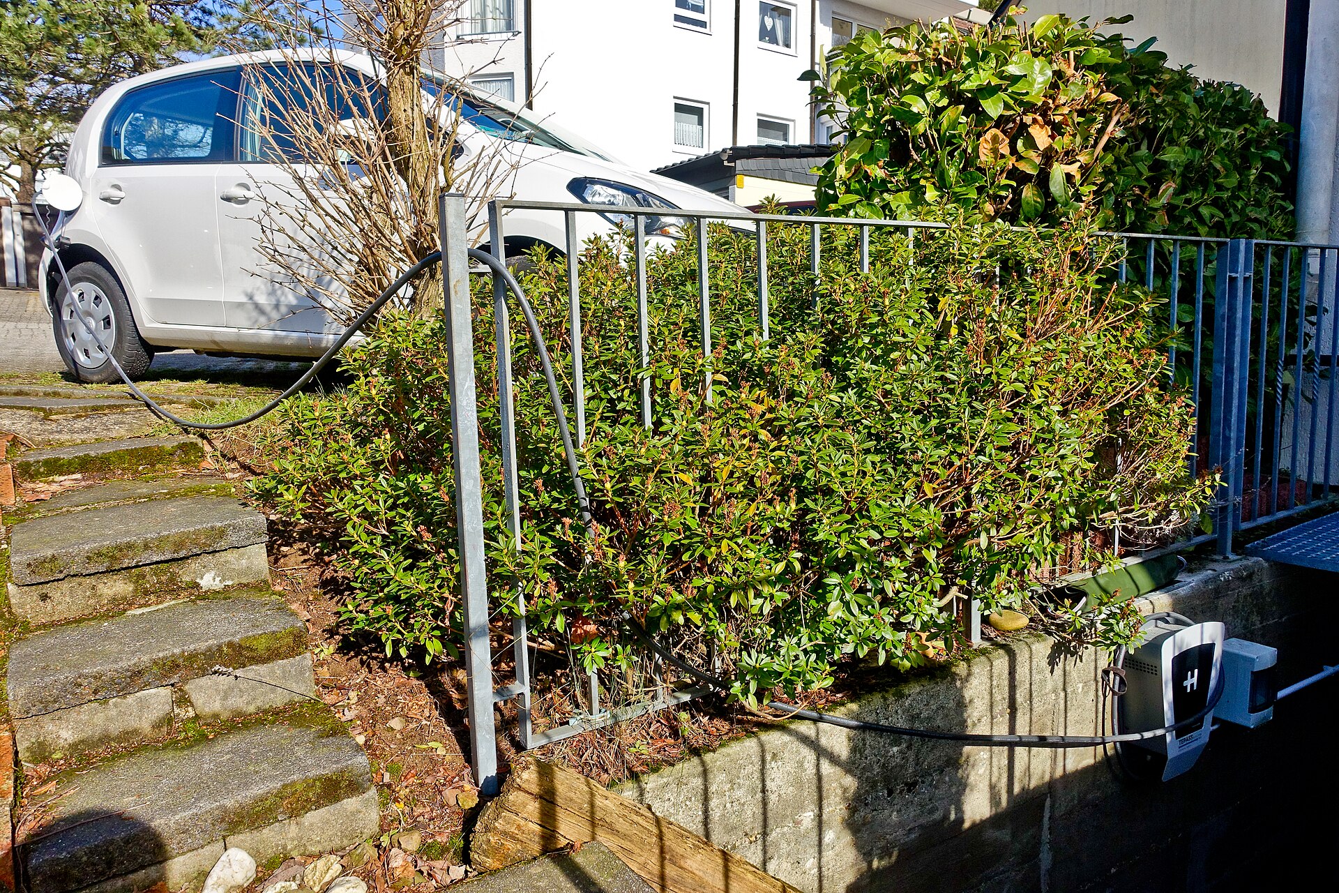 A small white electric car charging from a wall-mounted home charger, cable running across a garden wall.