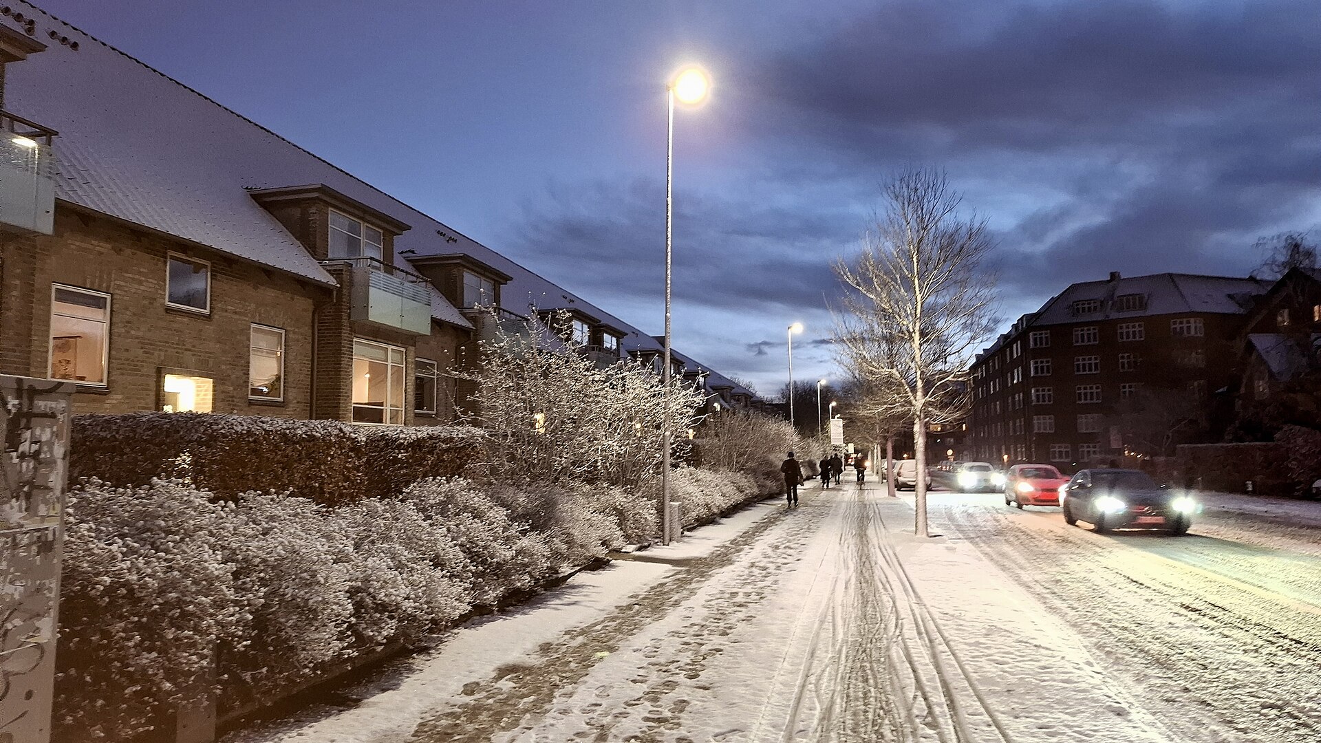 Cars with headlights on driving along a snow-covered European city street at dusk.