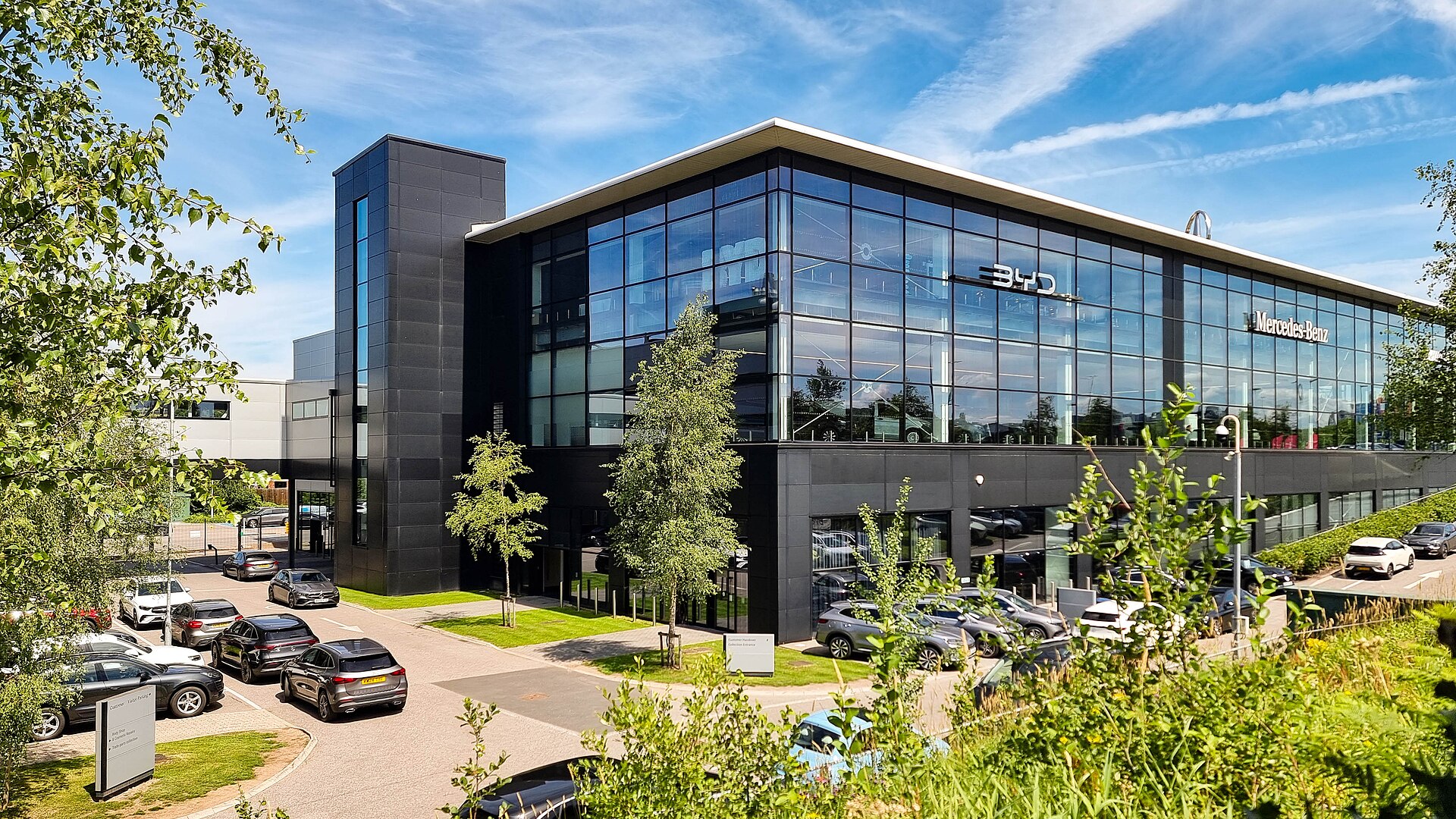 A glass-fronted multi-brand car dealership with new cars parked outside under a bright sky.